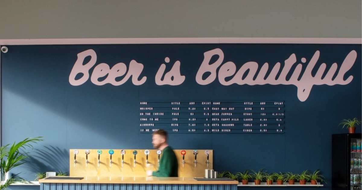 Inside Floc's taproom, the wall is painted with the cursive pink words 'beer is beautiful' and a person is visible behind the bar. The image conveys the welcoming and fun atmosphere of Floc's taproom, as well as the company's appreciation for the art of brewing